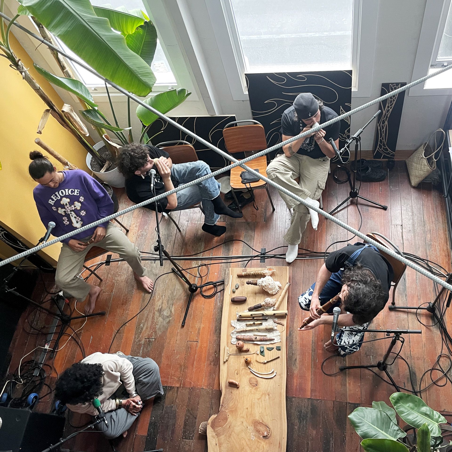 A birds-eye-view of a group of people sitting around a table in a well-lit room. Several taonga puoro are on the table, and the people are interacting with them. There are plants, and sunshine is coming through the window.